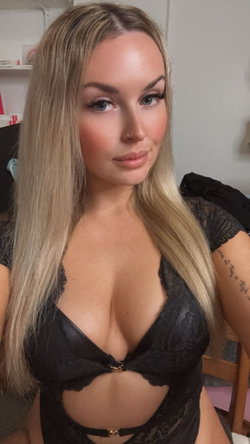 Blonde woman with long hair in black lace lingerie, posing indoors with visible arm tattoo.