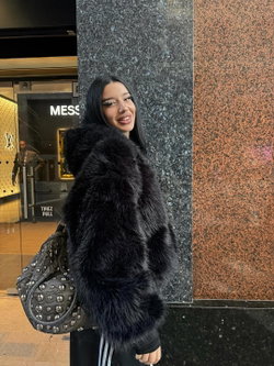A woman with long dark hair and bold makeup wears a black faux fur coat and carries a studded bag outside a luxury