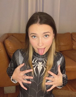 A woman with long brown hair and blue eyes sticks her tongue out while wearing a black spider-themed bodysuit, sitting