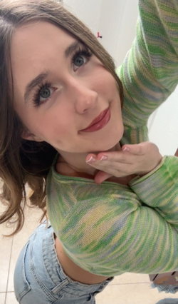 A young woman with brown hair and blue eyes wears a green striped crop top and denim shorts, posing in a selfie with