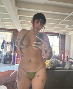 Curvy woman with dark hair and tattoos takes mirror selfie in olive green bikini, indoors with exposed beams and brick