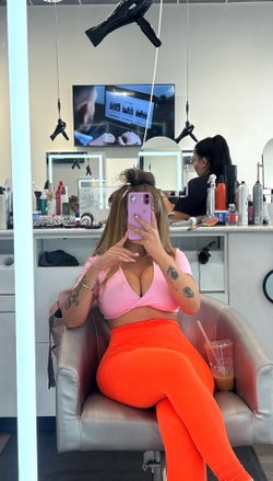 Curvy woman in pink crop top and orange leggings takes mirror selfie in salon, tattoos visible on arms.
