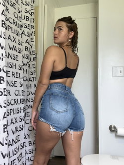 Curvy woman in black bra and denim cutoff shorts poses in a bathroom with a patterned shower curtain, looking over her