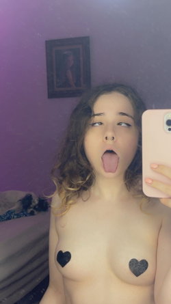 A young woman with curly hair takes a mirror selfie in a purple room, sticking her tongue out while bare-chested with