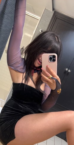 Brunette woman in a black velvet mini dress and sheer long sleeves takes a mirror selfie in a bathroom, smiling with