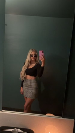 Blonde woman in black crop top and plaid mini skirt takes mirror selfie with pink phone, wearing sunglasses indoors.