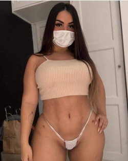 A woman with long dark hair wears a white face mask, a beige ribbed crop top, and white thong underwear, posing indoors