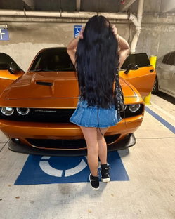 A woman with long dark hair stands back to camera in a denim mini skirt, posing in front of an orange Dodge Challenger