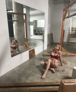 A woman with short red hair sits bound in rope on a concrete floor in a studio space, wearing a red top and black