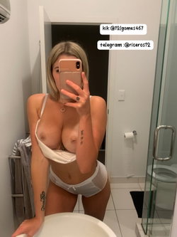 Blonde woman with curvy build takes mirror selfie in bathroom, wearing white lingerie with bare breasts visible,