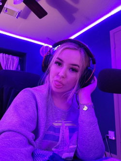 Blonde woman in a gray sweatshirt and headphones poses playfully in a room with purple LED lighting, making a puckered