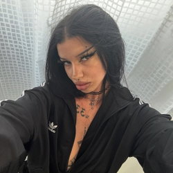 Brunette woman with dark hair and tattoos on chest, wearing black Adidas jacket, taking selfie in front of sheer