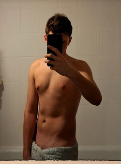 Shirtless man with short dark hair taking mirror selfie in bathroom, wearing towel around waist.