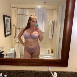 A curvy woman with blonde hair takes a mirror selfie in a lavender lace bra and matching underwear in a bathroom.