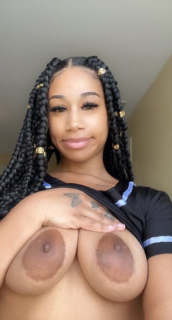 A woman with long black braids and a butterfly tattoo on her hand poses topless, holding her shirt down to reveal her