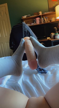 Person in gray socks holding a flesh-toned dildo between their feet on a bed, with a desk and green wall in the