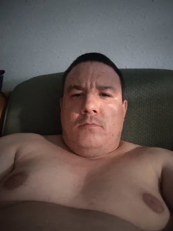 Shirtless man with short dark hair lying on a green couch, facing the camera in a dimly lit room.