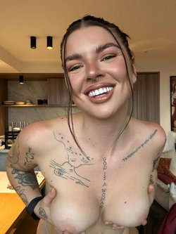 A person with tattoos and a nose ring smiles in a kitchen.