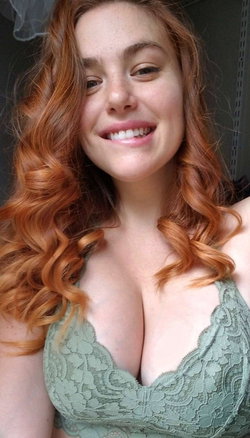 Redheaded woman with curly hair smiles in a green lace bra, indoors.