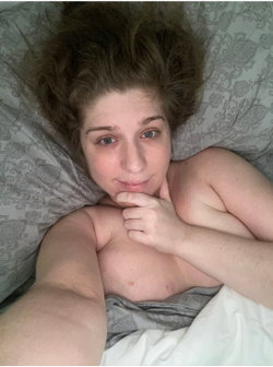 A woman with curly brown hair lies on a patterned gray pillow, bare-chested and partially covered by sheets, looking