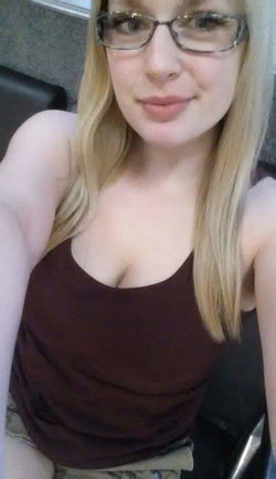 Blonde woman with glasses wearing a dark brown tank top, taking a close-up selfie indoors.