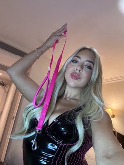 Blonde woman in a shiny black corset with pink trim holds a pink leash, posing for a selfie indoors.