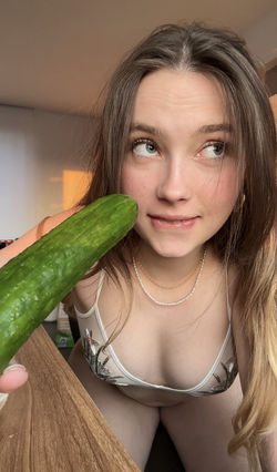 A young woman with long brown hair and blue eyes holds a cucumber near her face, wearing a sheer floral top indoors.