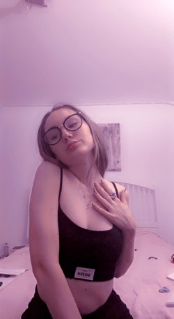 A woman with shoulder-length hair and round glasses poses in a black crop top, hand on chest, in a softly lit bedroom.