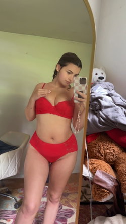 A woman with dark hair in red lace lingerie takes a mirror selfie in a bedroom with stuffed animals and laundry visible.