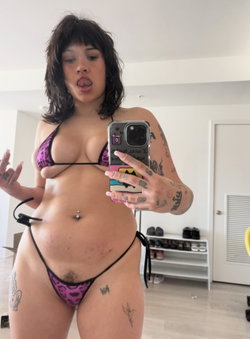 Curvy woman with dark hair and tattoos takes a mirror selfie in a purple patterned bikini, sticking out her tongue