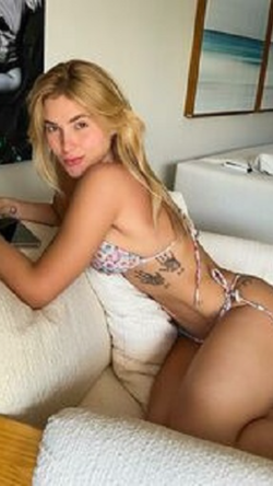 Blonde woman in floral bikini lounging on a white couch, showing side profile with visible tattoos on her back and hip.
