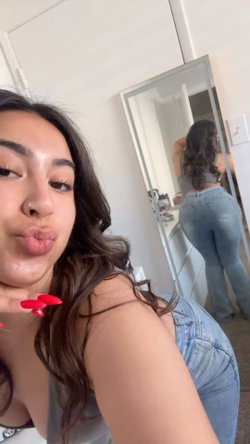 Curvy brunette woman with long hair takes a selfie in a gray tank top and jeans.