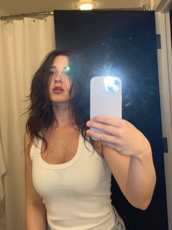 Woman with long dark hair in a white top taking a selfie in a bathroom.