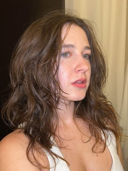 A brunette woman with wavy hair and a white top looks off-camera.
