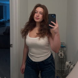 Curvy woman with long brown hair takes a mirror selfie in a white tank top and jeans, standing in a bathroom.
