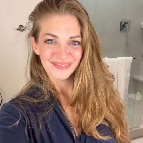 Blonde woman with long hair smiles in a bathroom selfie, wearing a dark blue robe.