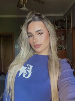 A woman with long blonde hair wearing a blue sweater takes a selfie in a living room.