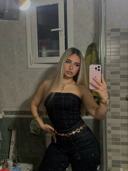 Blonde woman in a black strapless top and jeans taking a selfie in a tiled bathroom.