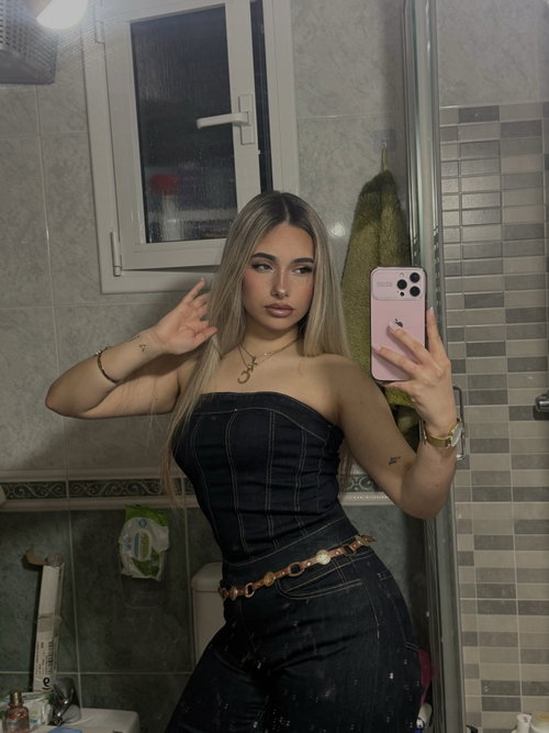 Slim curvy blonde takes a selfie in bathroom wearing strapless denim top and jeans.