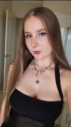 A woman with long brown hair and a nose ring wears a black halter top and layered necklaces, posing indoors in a