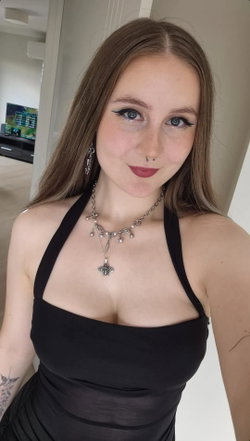 A woman with long brown hair and makeup wears a black halter top, layered necklaces, and has visible tattoos on her