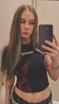 A young woman with long light brown hair takes a mirror selfie, wearing a black crop top with red dragon design and