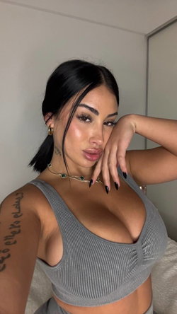 Brunette woman with dark hair in a low ponytail, wearing a gray ribbed crop top, posing indoors with hand on cheek and