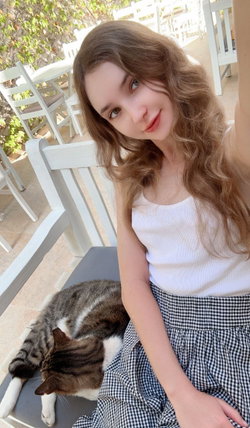 A young woman with long wavy brown hair sits on a white chair outdoors, wearing a white tank top and checkered skirt,