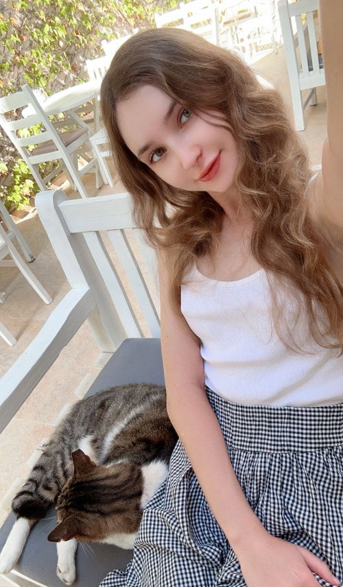 A young woman with long wavy brown hair sits on a white chair outdoors, wearing a white tank top and checkered skirt,