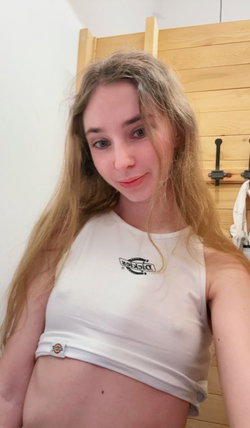 A blonde woman with long hair wears a white cropped tank top, standing indoors near a wooden headboard, smiling at the