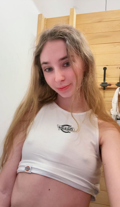 A blonde woman with long hair wears a white cropped tank top, standing indoors near a wooden headboard, smiling at the