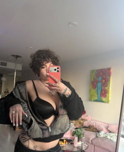 Curly-haired person in a black outfit taking a selfie in a living room.