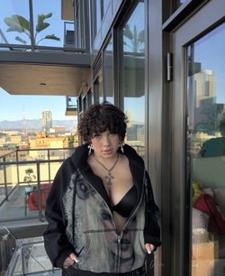 Person with curly hair in a sheer top and open jacket stands by a window overlooking a city.