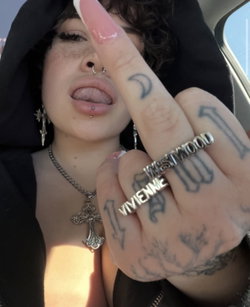 A person with dark hair and tattoos poses in a car, wearing a cross necklace and earrings.